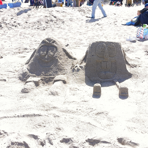 Das Bild zeigt zwei Sandfiguren auf einem Strand, die nach bekannten Figuren aus einem Kinder-Fernsehprogramm geformt wurden. Die Figuren sitzen nebeneinander und haben ausdrucksstarke Gesichter mit großen Augen und breiten Lächeln. Die Sandfiguren sind detailliert gestaltet, mit sichtbaren Gesichtszügen und Beinen, die aus dem Sand herausragen. Im Hintergrund sind weitere Menschen zu sehen, die sich auf dem Strand befinden, einige sitzen und andere stehen. Es gibt auch einige Gegenstände wie Kisten und ein Kinderwagen, was auf eine Familienaktivität hinweist. Die Sonne scheint, und der Sand ist weiß und fein.