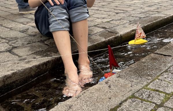 Kopfsteinpflaster mit einer schmalen Rinne in der Wasser fließt. Eine Person sitzt an der Rinne und hat seine Füße im Wasser. Neben den Füßen schwimmen zwei Holzboote mit bunten Segeln. Der Oberkörper der Person ist nicht zu sehen.