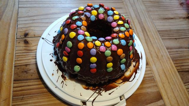 Marmorkuchen in Guglhupfform mit Schokolade überzogen und mit Smarties besetzt. Der Kuchen steht auf einer weißen Kuchenplatte, die ihrerseits auf einen Holztisch steht.