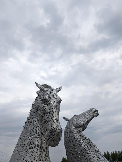 Two enormous metal sculptures of horse heads at the bottom of the frame, one facing towards camera with ears back and the other rearing back and facing off to the right. The cloudy sky above fills the top half of the frame.