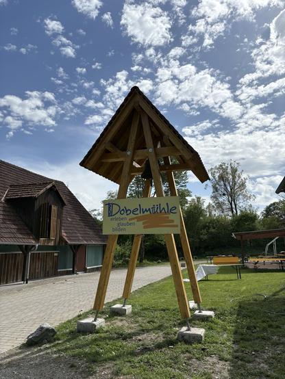 Ein Glockenturm aus Balken mit kleinem Dach, mit dem Schild davor: Dobelmühle - erleben, glauben - bilden. Im Hintergrund ein altes Stallgebäude.