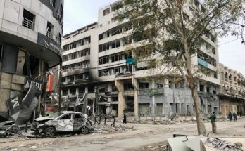 L'immagine mostra una strada e dei palazzi di gaza gravemente distrutti dai bombardamenti. Anche la strada e un'automobile parcheggiata sono gravemente danneggiate.