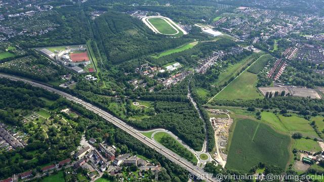 An aerial photo of Gelsenkirchen. There is a freeway (the A42) in the front. You can see the half round building of the Gelsenkirchen Prison in the top left. In the top distance you can see the harness racing track. There are many trees and grass areas around and some living quaters.