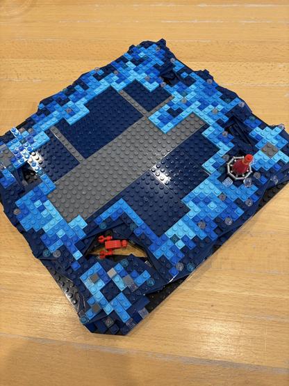 Eine Bluebrixx-Kreation mit verschiedenen Blautönen, die Wasser und Wellen darstellen. Das Design umfasst eine graue Plattform in der Mitte und kleine rote Elemente, die einen Krebs und eine Boje darstellen. Es ist die Basisplatte für den Leuchturm des Astronomen.
