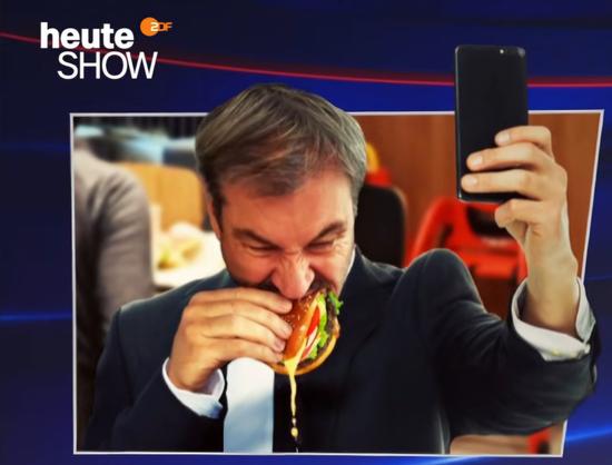 Markus Söder isst einen Hamburger und verzieht dabei sein Gesicht. Käse tritt seitlich heraus und tropft hinunter. 
Mit der linken Hand macht er ein Selfie.