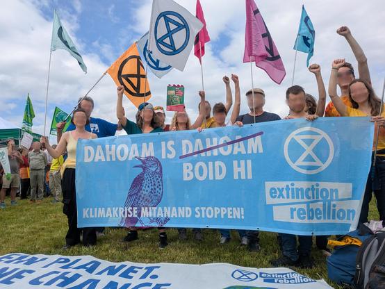 XR Protest in Reichling bei der KoaGas Demo. XR Aktivisti hinter einem großen Banner  mit XR Logo u. Vogel sowie Schriftzug"Dahom is -Dahoam- Boid Hi" (Dahoam durchgestrichen) und "Klimakrise mitanand stoppen!"