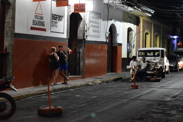Dark city street in Leon Nicaragua, two people leaning on left against hostel wall. Two people sitting on right on bumper of old truck. Harsh white light illuminates the street.