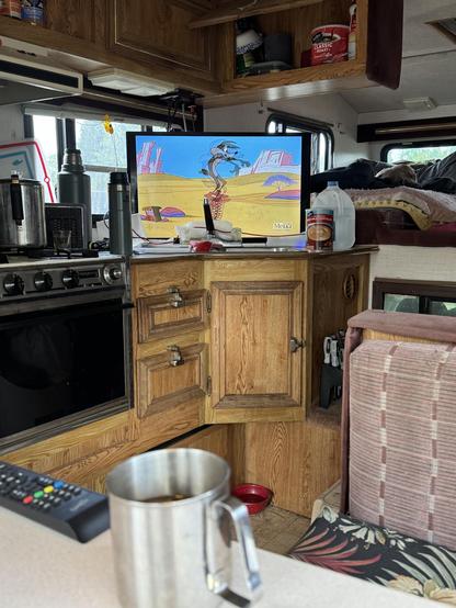 An interior view of a camper van features a wooden kitchen area with a coffee mug in the foreground. A television displays a cartoon scene, while various kitchen items and a sleeping dog are visible in the background.