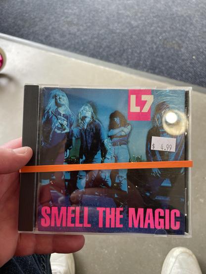Image of a CD titled "Smell the Magic" by the band L7. The cover features the band members in a colorful, blue-toned photograph with bold pink text. The CD is held in a hand and has a price tag of $4.99