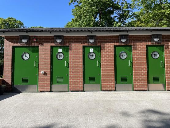Row of five modern public toilets in a park of red brick construction with green doors. Each toilet has a sign on it to show that men and women can use it. The toilet on the far left has a bigger door as it is an accessible one.