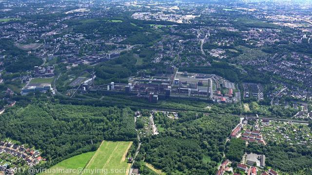 An aerial photo of the UNESCO world heritage site Zollverein. In the middle of the image is the coking plant Zollverein in the full view. The coal mine Zollverein can be seen in the top left of it. It is sourrounded by green parks, woods/trees and living/working quaters.