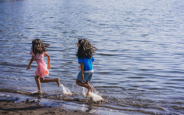 The image shows two children playing at the edge of a body of water. They are running through the shallow water near the shore, with one wearing a pink outfit and the other in a blue shirt and shorts. The scene appears joyful and lively.