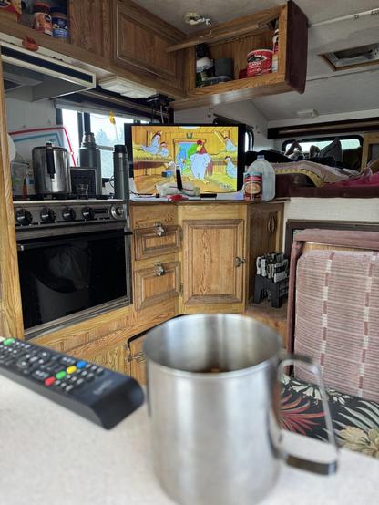 An interior view of a camper featuring a wooden kitchen area with a stove and shelves holding various food items. A television displays a cartoon with animated chickens. In the foreground, there is a metal cup on a countertop and a remote control nearby.