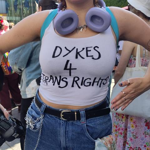 A woman wearing a shirt saying "Dykes for trans rights" She is also wearing jeans with a leather belt and chains and a pair of purple headphones around her neck