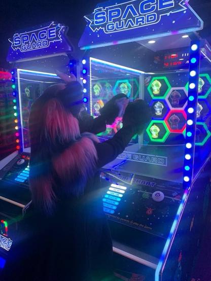 Noir (Nirik (Kirin) fursuit with white, pink and blue mane) plays a space guard arcade game.