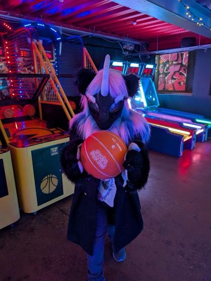 Noir (Nirik (Kirin) fursuit with white, pink and blue mane) holds a basketball in front of a basketball arcade game.