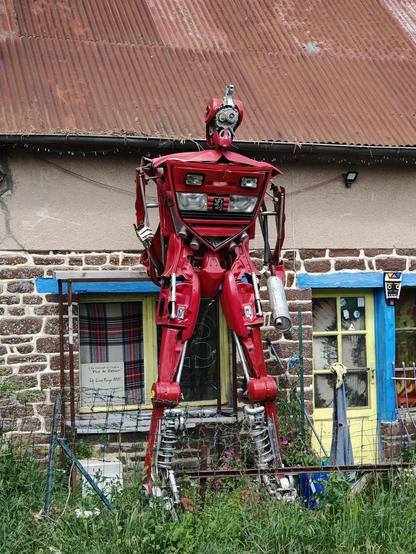 Devant une modeste maison normande, à la toiture en tôles ondulées rouillées, un grand totem/robot rouge constitué de diverses pièces de Peugeot 205 🚗