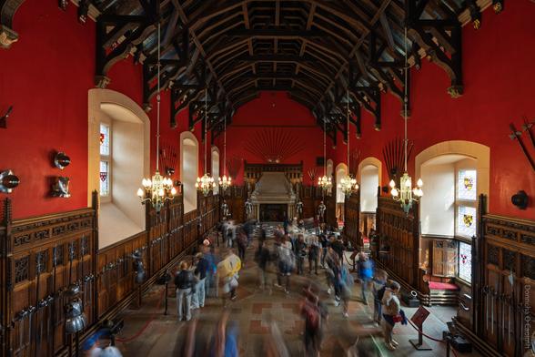 Wide-angle interior photograph of The Great Hall at Edinburgh Castle in Scotland, featuring a high vaulted wooden ceiling, red walls adorned with medieval armour and weapons, stained glass windows, ornate chandeliers, and a large stone fireplace at the far end, with a blurred crowd of visitors moving through the hall.