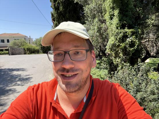 Me, wearing a green baseball hat and orange polo shirt, in the burning sun. Some shrubs and buildings behind me.