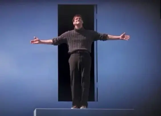 A man in black t-shirt and trousers standing happily with stretched arms and eyes closed in front of an enclosure that is painted in blue. It's a scene from "Truman Show" movie and the man is Jim Carry.