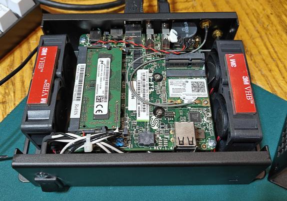 The image shows the internal components of a small, open computer case. Here's a breakdown of what's visible:

* **Motherboard:** A green printed circuit board (PCB) dominates the center, which is the motherboard. Various electronic components, chips, and connectors are soldered onto it. You can see labels like "Bt-P13" and "HDMI" printed on it.
* **RAM:** To the left of the motherboard, a stick of green RAM (Random Access Memory) is inserted into a slot. It has a white label with barcodes and technical specifications.
* **Wireless Card:** To the right of the RAM, a smaller green PCB with a white label appears to be a wireless network card. Two antenna cables are connected to it.
* **Cooling Fans:** On both the left and right sides of the case, black cooling fans are mounted. They have red 3M VHB™ tape securing them.
* **Connectors:** Various connectors are visible on the motherboard, including what appears to be an HDMI port, USB ports, and power connectors.
* **Cables:** Several colored wires and cables are routed within the case, connecting different components. Some are bundled with white zip ties.
* **Case:** The components are housed in a black metal case that is currently open, revealing the internal hardware.
* **Background:** The computer case is sitting on a surface that appears to be a green mat or desk pad.

Overall, the image provides a detailed view of the internal layout and components of a compact computer system.