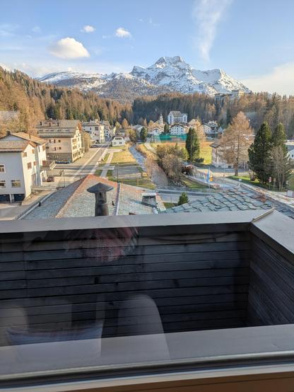 View above and over the town of Sils. A small road on the left cuts through the town, buildings either side in very alpine style (pointed roofs etc) and to the right, a bubbling, clear stream winds through the town. In the background, a snow capped peak