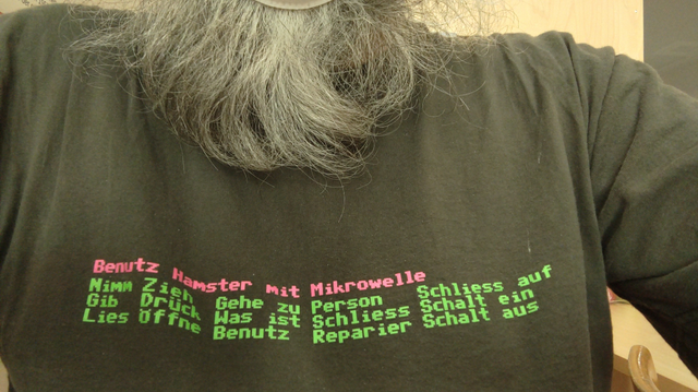 Selfie of my dark grey t-shirt and silvergrey, curly beard.

The t-shirt reads (in German):

In magenta: Use Hamster with Microwave

In Green:

Take  Pull  Go to  Person  Unlock
Give  Push  What is  Lock  Turn on
Read  Open  Use  Fix  Turn off