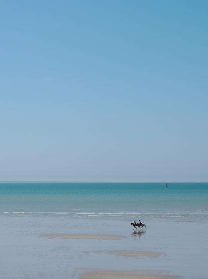 Deux chevaux et leurs cavaliers, en train de trotter sur la plage de Kairon en Normandie, la mer est calme, le ciel bleu sans nuage. On aperçoit, au loin, les îles Chausey et deux petits voiliers dans les eaux turquoises.