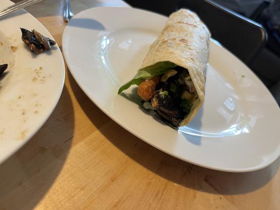 Ein Foto von einem eingerollten Shawarm, von der Seite. Das gerollte Fladenbrot ist seitlich offen und man sieht einen sehr dunkles Stück Portobello, Salat, einen Röstistick und ein Stück Gurke.