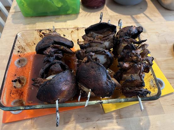 Ein Foto von den Pilzspießen wie sie sehr dunkel aus dem Ofen kommen. Der Boden der Auflaufform ist mit einem dunklen Fond bedeckt. Auf den Rändern liegen 3 Metallspieße bestückt mit Portobelle, Austernpilzen und Zwiebeln.