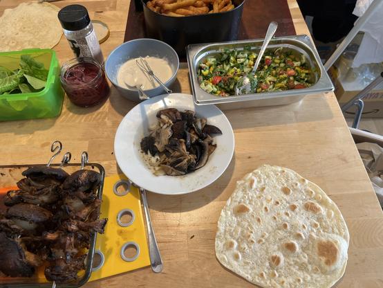 Ein Foto vom Tisch mit dem "Baukasten". Oben eine Schüssel mit gerupften Salatblättern, ein Glas mit eingelegten roten Zwiebeln, eine Schale mit einer beigen Tahini-Joghurt-Sauce und GN-Behälter mit klein gewürfeltem Gemüse (gelbe Paprika, Tomaten, Gurke, viel Petersilie). In der Mitte ein tiefer Teller mit vom Spieß abgeschnittenen Pilzen. Unten nochmal 2 Spieße über der Auflaufform und ein frisch gemachtes Fladenbrot, das gleich belegt wird.