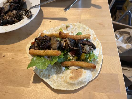 Ein Foto von einem belegten noch nicht gerollten Shawarma. Erste Schicht Salatblätter, dann Joghurtsauce, Gemüse, Pilze und links und rechts Rösti-Fries.