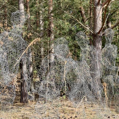 Mehrere aus Maschendraht geformte Menschenfiguren in unterschiedlichen Haltungen vor einer Reihe von Nadelbäumen. Während die Köpfe und Arme recht gut zu erkennen sind, verschmilzt der Rest der Figuren großenteils mit der Umgebung.