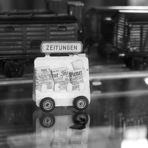 Das Model von einem alten, weißen Zeitungswage mit drei unterschiedlichen Zeitungen vor einer Märklinbahn. Alles klein aber trotzdem super Darstellung. Das Foto ist in Schwarzweiß.