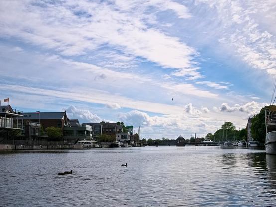 In the foreground, the water of a harbour. Buildings and boats to the left and right. A dark bridge spanning the water can be seen on the horizon. There are some ducks on the water, a bird (probably a seagull) flies in the sky. And above it a lot of sky with white streaks of cloud, scattered fluffy clouds and patches of blue.