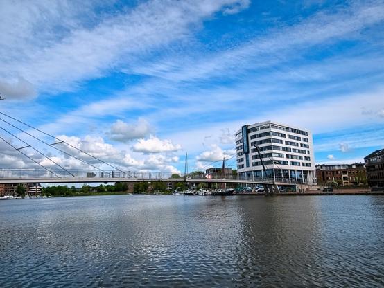 In the foreground, the water of a harbour. A curved bridge spans the water. A box-shaped white tower block can be seen on the other bank. The sky is mainly blue with white streaks of cloud and individual fluffy clouds.