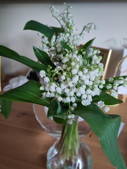 des fleurs blanches en clochette (muguet) dans un vase, avec quelques feuilles vertes. le tout est posé sur une table en bois.