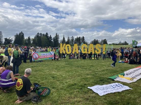 Menschen stehen auf einer Wiese im Kreis. Sie haben Banner dabei und halten gelbe Buchstaben hoch die den Text „Koa Gas!“ bilden