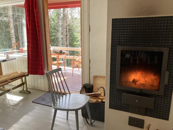 Interior view of a cabin in sunlight. On the left a doorway to a porch. On the right a whitewashed fireplace with flames visible through its glass hatch.