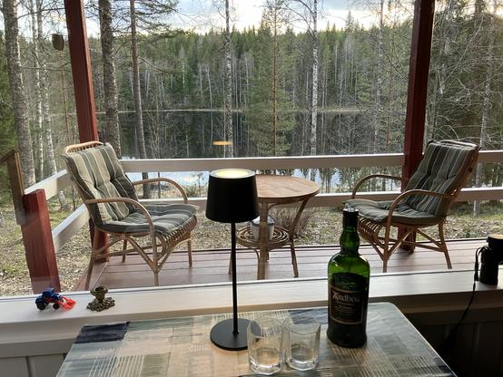 A bottle of Ardbeg Uigeadail and two glasses on a table by a large window. Beyond the window is a porch with wicker table and chairs, and beyond that a view to a lake surrounded by birch, pine and spruce forest.
