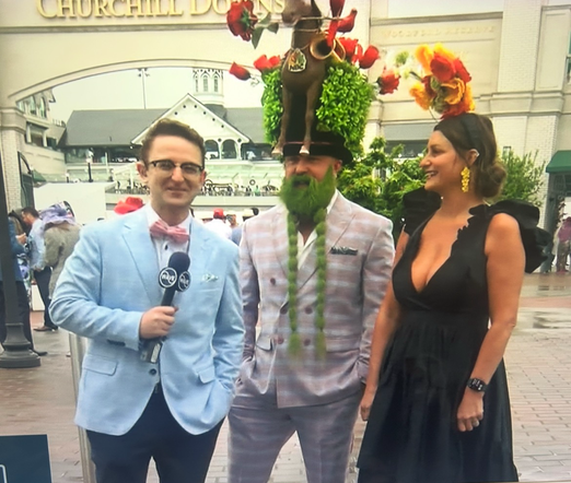 Image of male and female reporter interviewing a male guest to the Kentucky Derby. He’s wearing a large hat covered in greenery and a horse and roses. His beard is dyed green with long green braids. He is wearing a dapper laid suit.