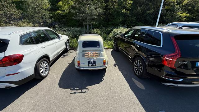 Ein beiger, originaler Fiat 500 der ersten Baureihe auf ejnem Parkplatz zwischen zwei aktuellen Kombis. Er sieht aus wie ein Spielzeugauto. Von hinten fotografiert.