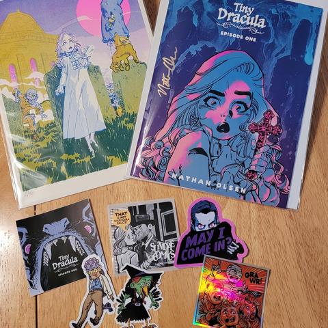 A print, a comic, and a bunch of stickers. The risograph print is on the left and is notable in person for its hot-pink moon. The comic cover features the main character, Lucy, clutching an ornate cross. One of the stickers has a shiny foil background.