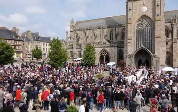 A large crowd gathers in front of St Tugdual Cathedral in Tréguier during the procession. People are dressed in various attire, and some are holding banners.