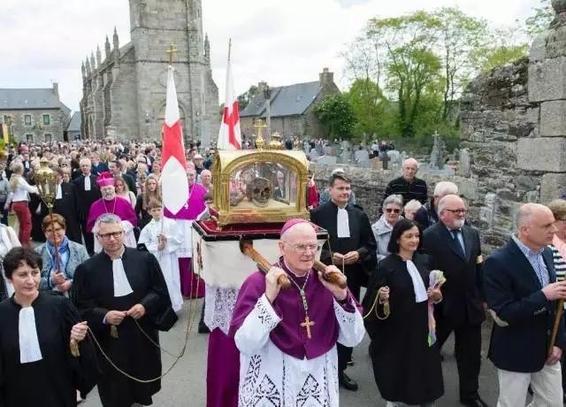 A procession featuring a large group of people, including clergy in liturgical attire and lay participants, walks through a stone street. They carry a religious relic in a gold-hued container. The background includes church and historical buildings in Minihy Tréguier, with trees and flags