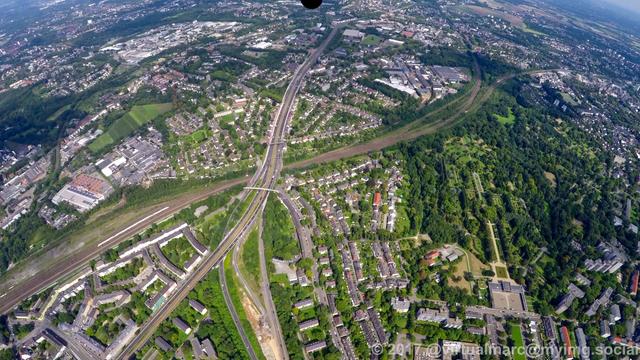An aerial photo of two freeways. The main freeway, the A40 is going through the image and crossing the main trainline over a bridge. The A52 is coming from the bottom middle and ends in the A40. There is a big park with trees on the right which is the park cementry.