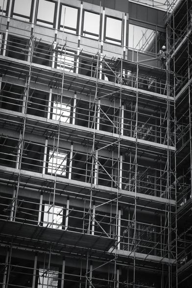 Black and white. A builder is standing on a wooden platform on scaffolding near the top of a building, looking down.