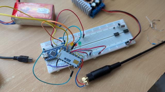A breadboard with an Arduino nano, SA818 module, a few LEDs, and lots of wires. There is a battery with a DC-DC converter above it, and a short whip antenna next to the board.