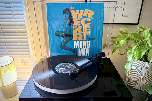 A black vinyl record spins on a black turntable next to a green houseplant. The record sleeve stands up in the turntable’s dust cover behind the platter.