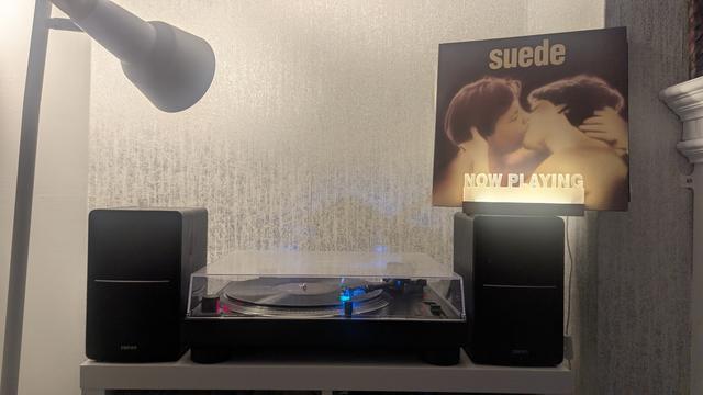 We have a black turntable flanked by two black speakers on some white furniture with grey wallpaper behind. The playing record is black vinyl. On the right speaker are the words 'Now Playing' lit up in front of the record sleeve.

The sleeve shows the naked head and shoulders of two white short-haired androgynous figures kissing passionately. There is a golden glow about them and the band's name is above this in lower case gold letters.